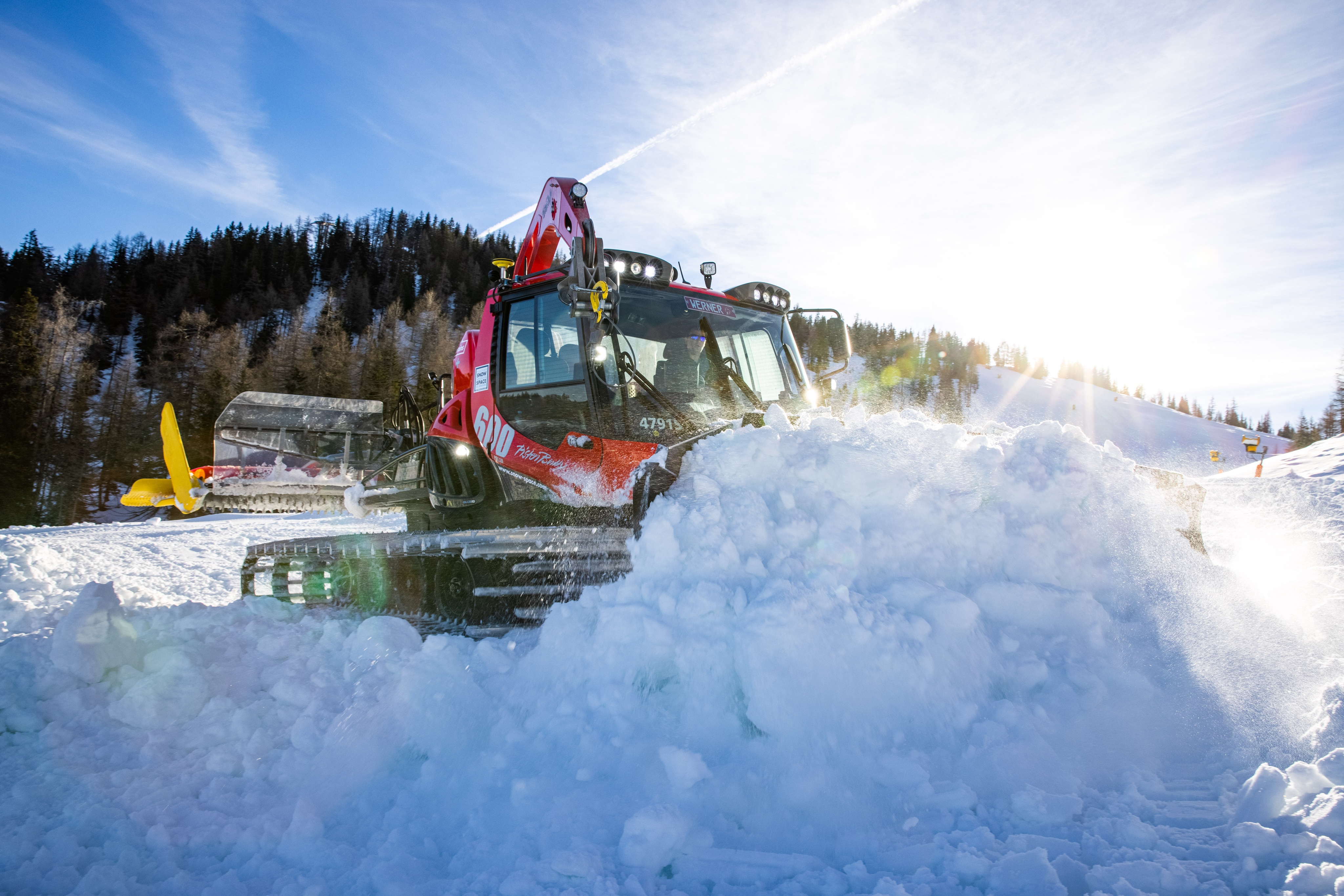 Pistenraupe schiebt einen Schneehaufen zusammen, Sonne über dem Wald